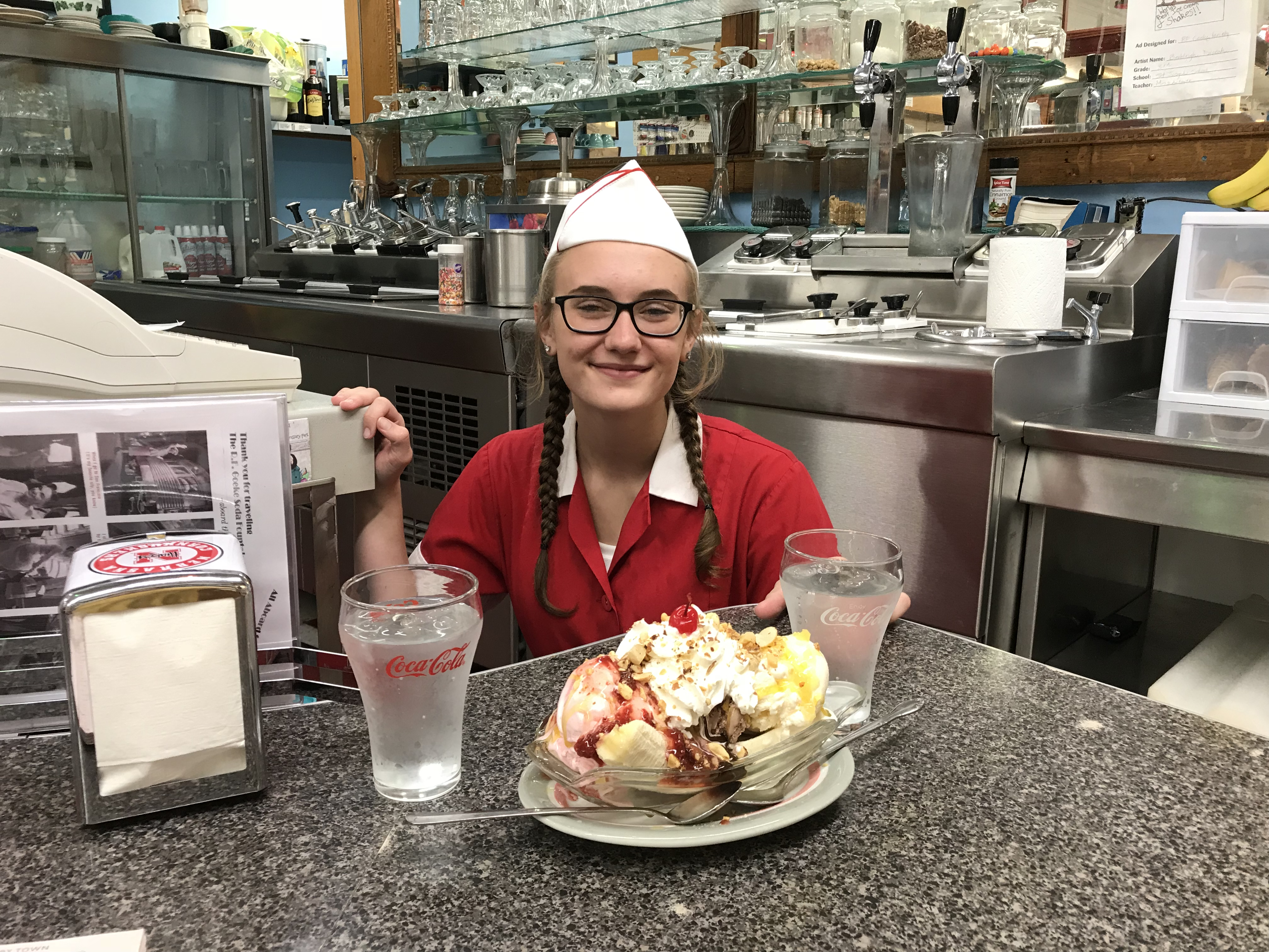 A woman stands behind a counter and presents a banana sundae piled with toppings and ice cream at R.F. Goeke Variety and Soda Fountain in Atkinson, Nebraska.