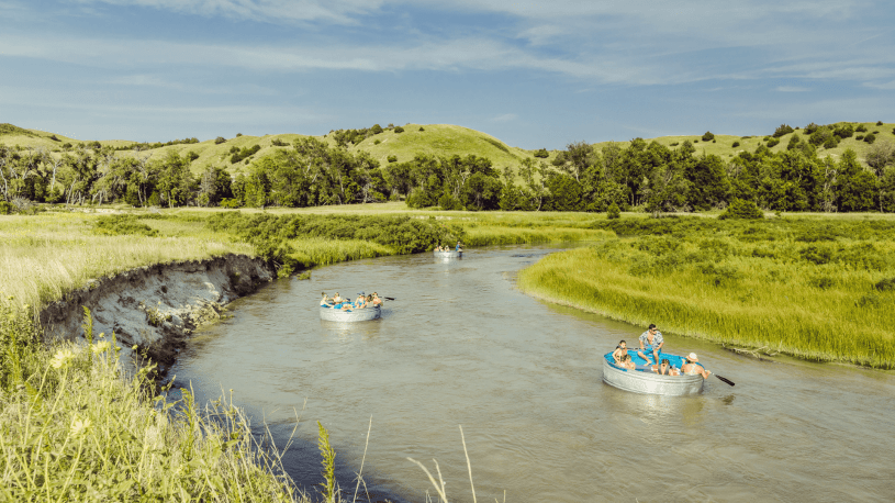 Two tanks floating down a Nebraska river with a scenic landscape of hills and grasslands surrounding the river.